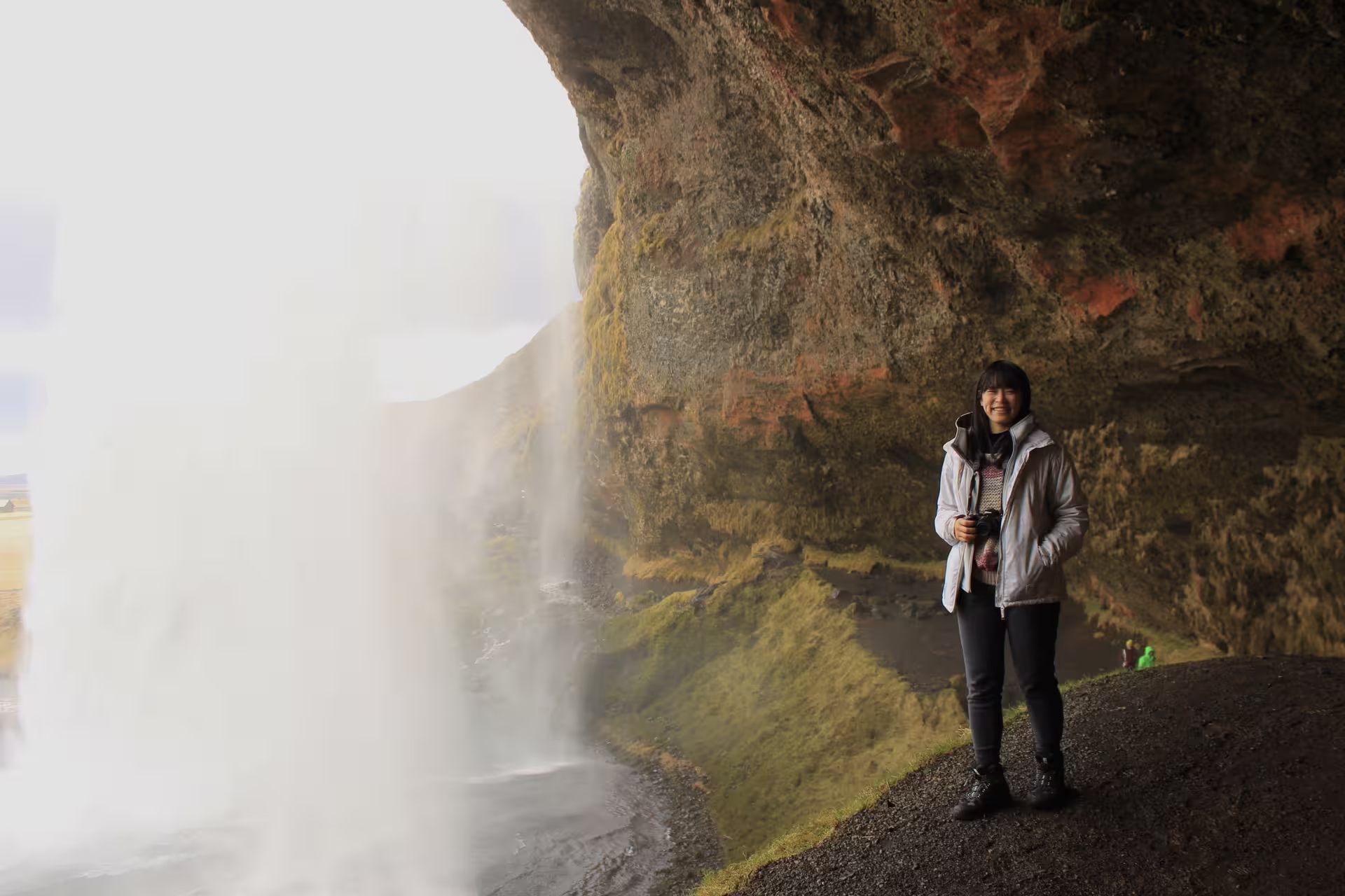 Smiling traveler standing behind Seljalandsfoss waterfall on Iceland's south coast holding a camera