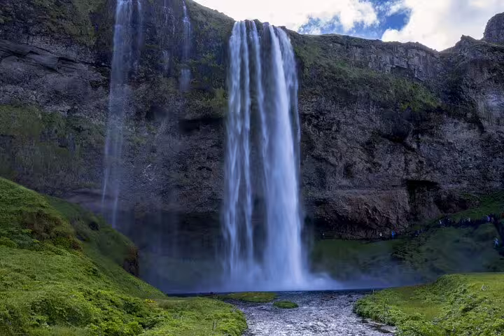 Tall waterfall cascading down mossy black basalt cliffs into a rocky stream on Iceland's South Coast