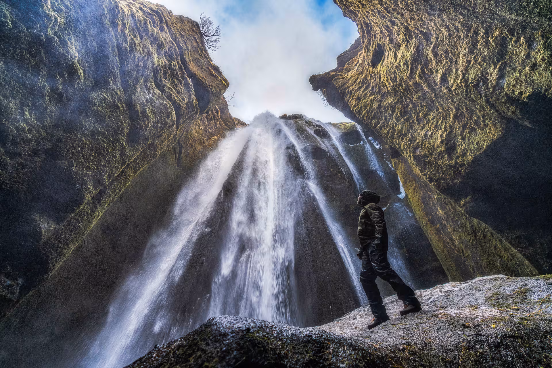 Person standing beneath a massive waterfall inside a rocky canyon on Iceland's South Coast
