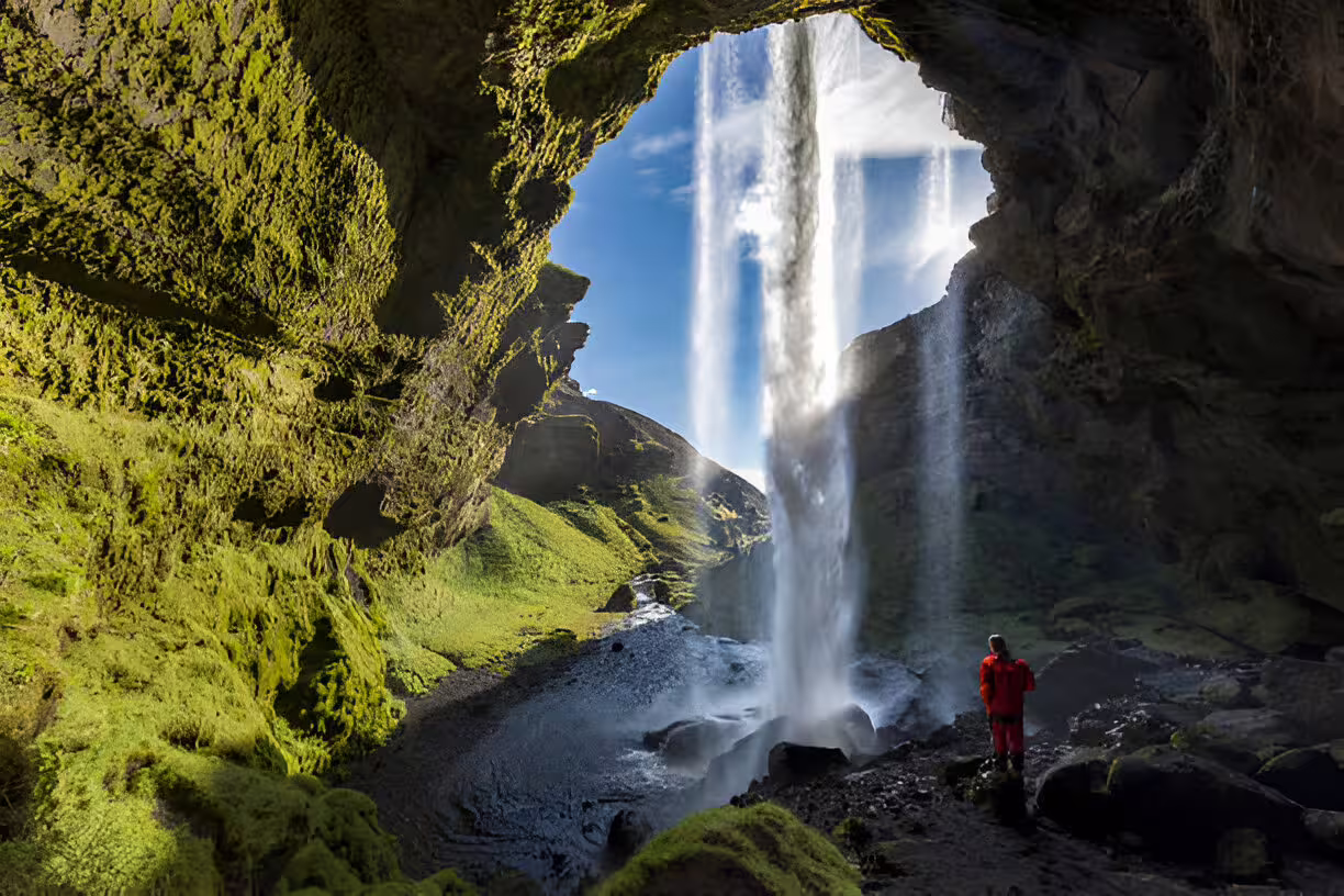Hiker in red jacket standing behind a massive waterfall viewed from inside a cave on Iceland's south coast