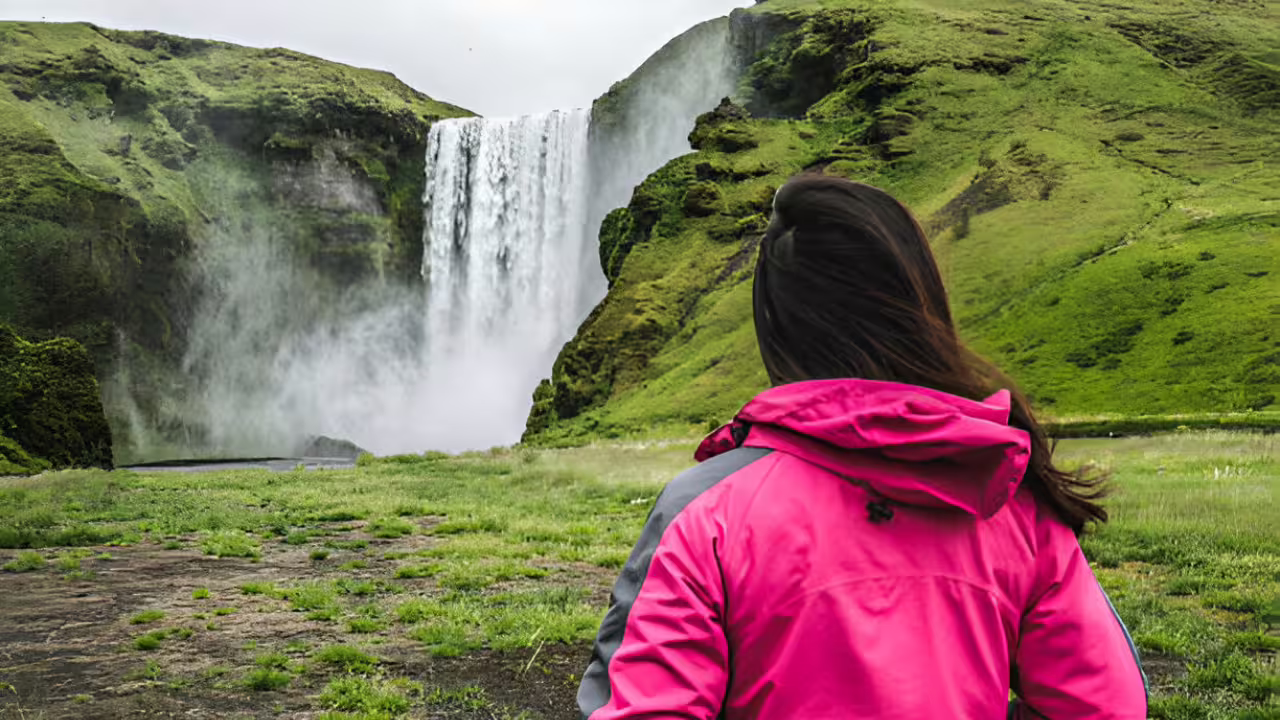 Woman in pink jacket gazing at a massive waterfall surrounded by green cliffs in Iceland