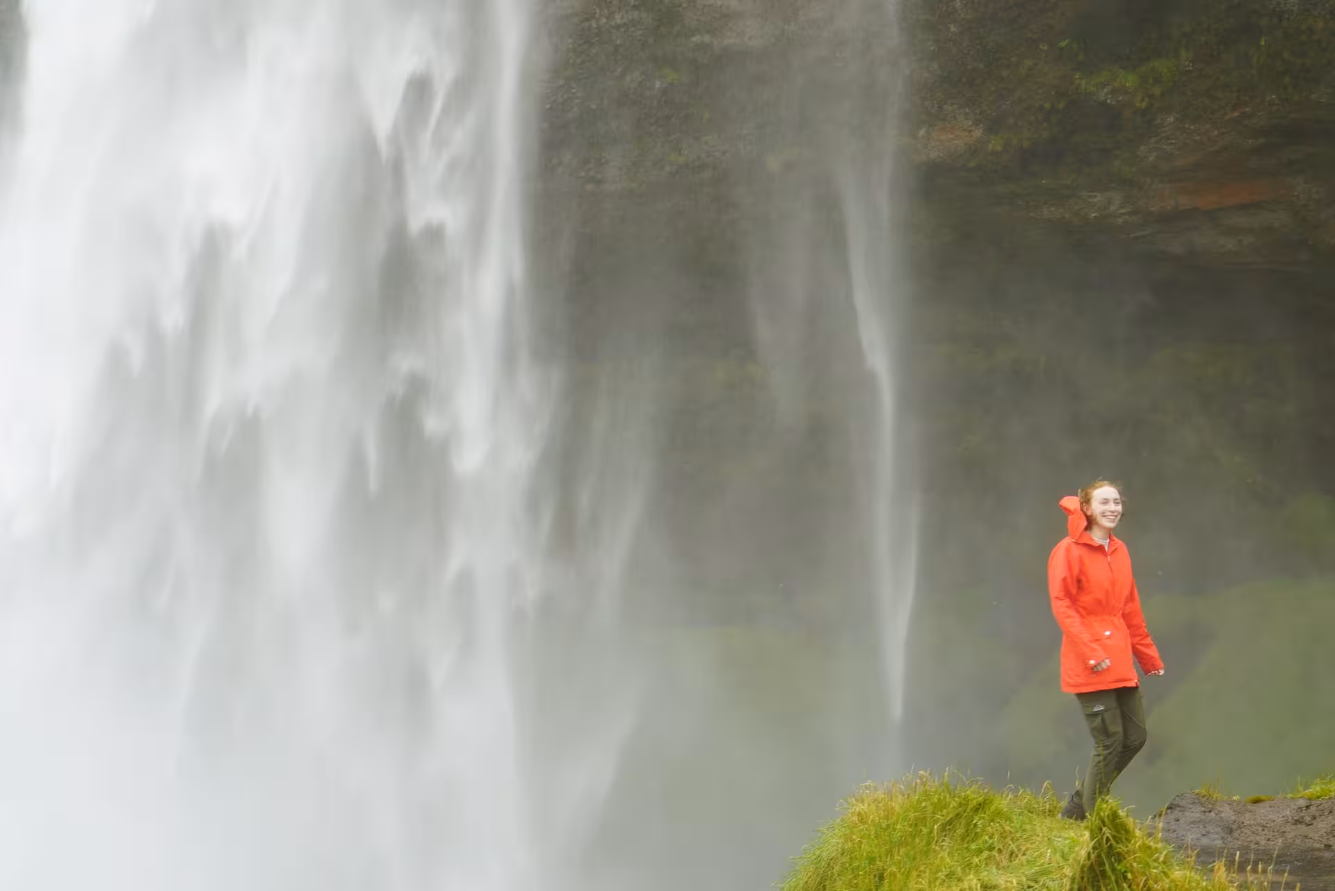 Woman in red jacket smiling beside massive Seljalandsfoss waterfall on Iceland's South Coast