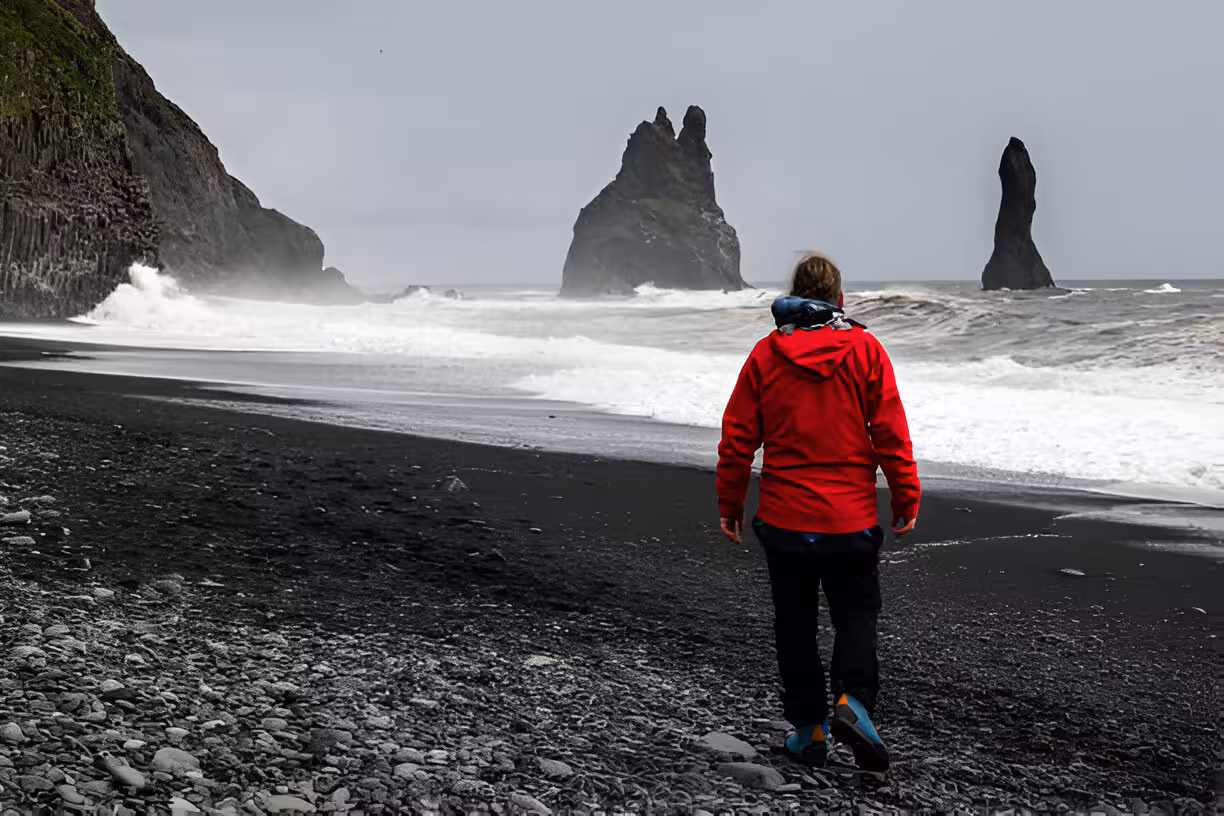 Person in red jacket walking on Iceland's black sand beach with dramatic basalt sea stacks in stormy waves