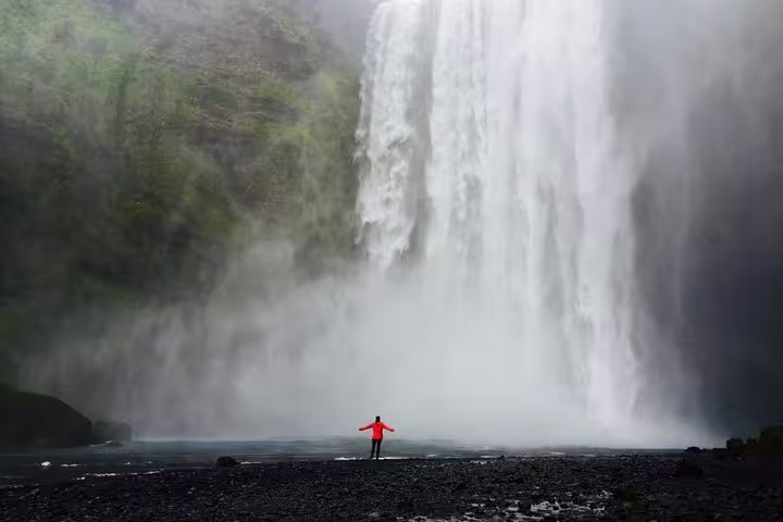 Person in red jacket standing before massive Skógafoss waterfall on Iceland's south coast