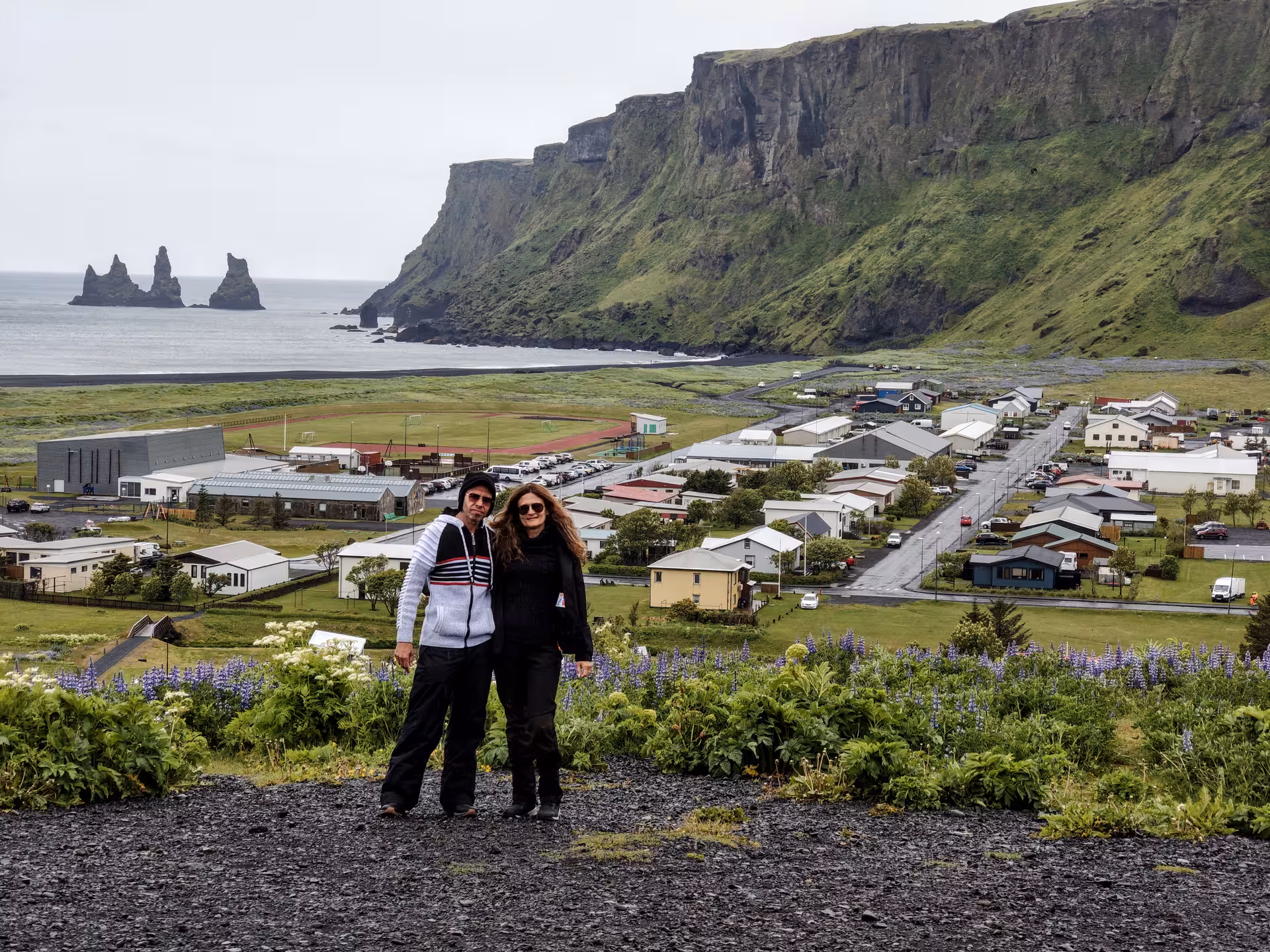 Couple posing above Vík village with black sand beach, sea stacks, and green cliffs on Iceland's South Coast