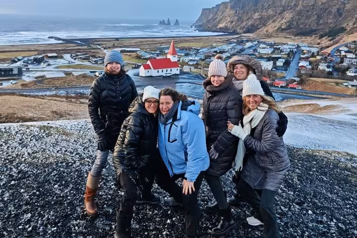 Group of six women posing on hilltop overlooking Vík village with red-roofed church and black sand beach, Iceland