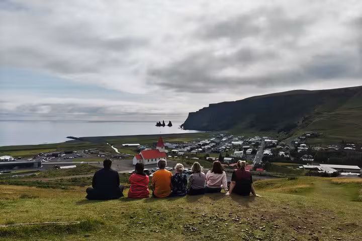 Group of tourists sitting on a hill overlooking Vík village, black sand beach, and coastal cliffs in South Iceland