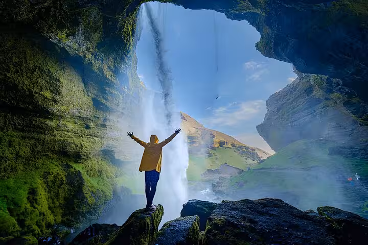 Person in yellow jacket standing behind waterfall with arms raised on Iceland South Coast tour