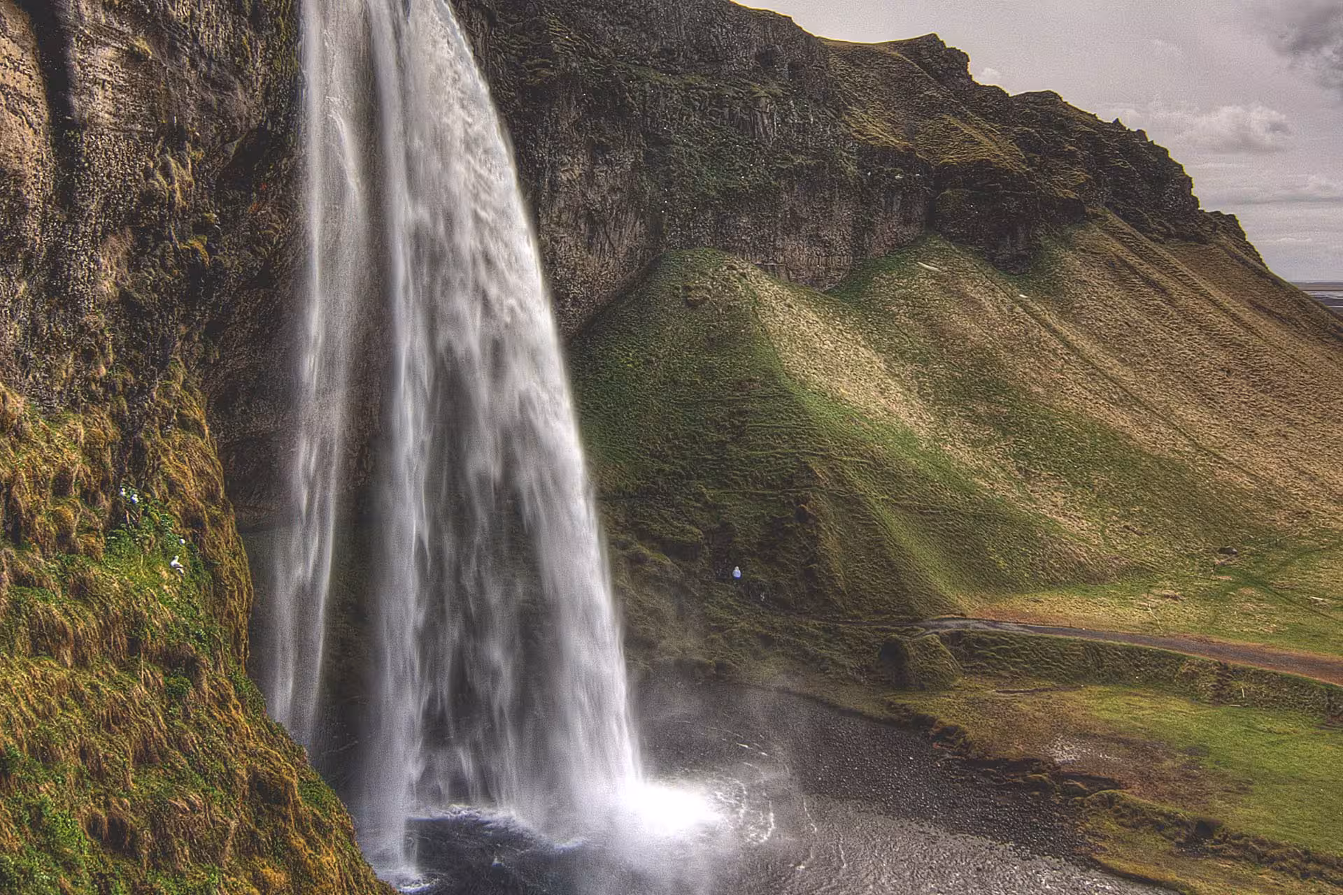 Massive waterfall cascading down mossy cliffs on Iceland's South Coast with green mountain backdrop