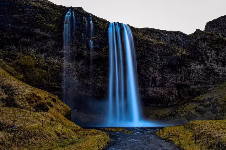 Tall blue waterfall cascading down dark volcanic cliffs into a river on Iceland's south coast