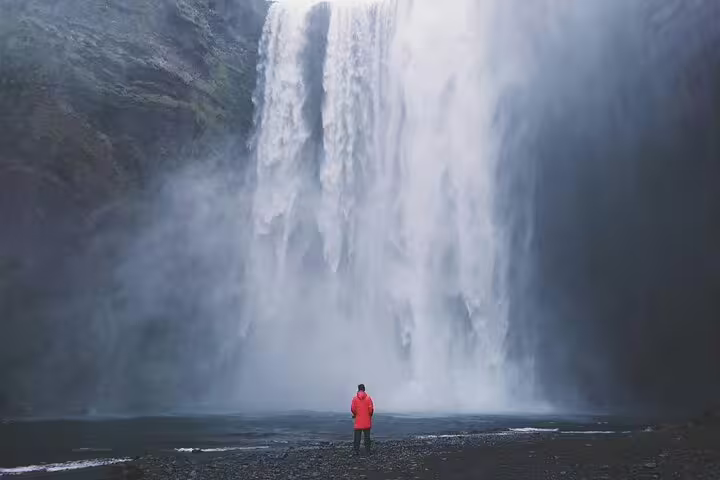 Person in red jacket standing before massive Skógafoss waterfall on Iceland's South Coast