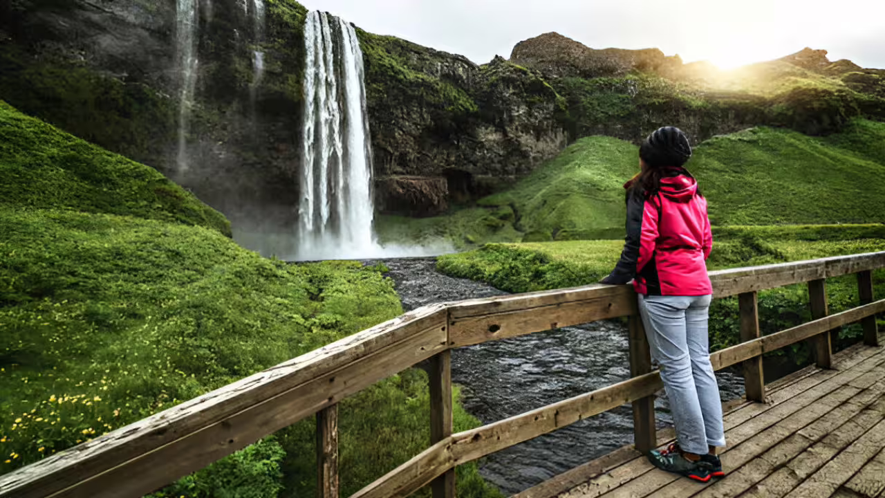 Hiker in red jacket standing on wooden bridge admiring Seljalandsfoss waterfall on Iceland's South Coast