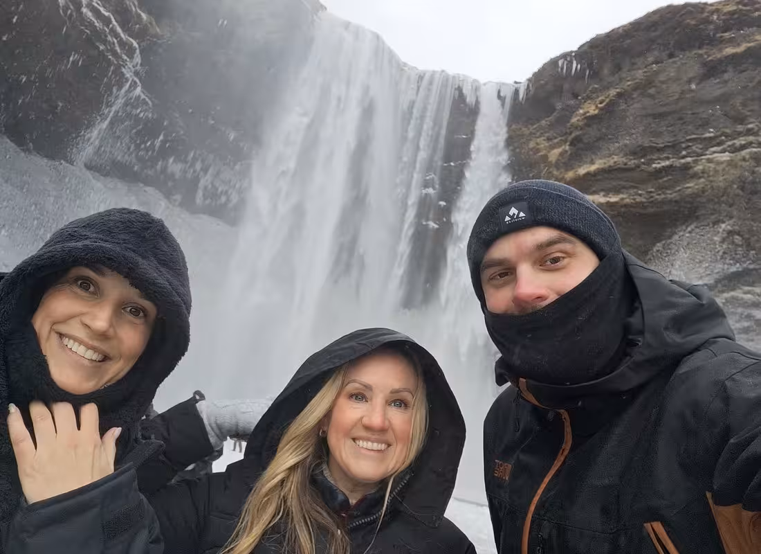 Three smiling tourists in winter gear posing in front of a large Icelandic waterfall on South Coast tour