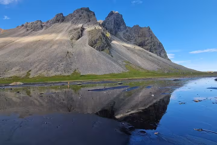Jagged volcanic mountain reflected in black sand beach water, South East Iceland under clear blue sky