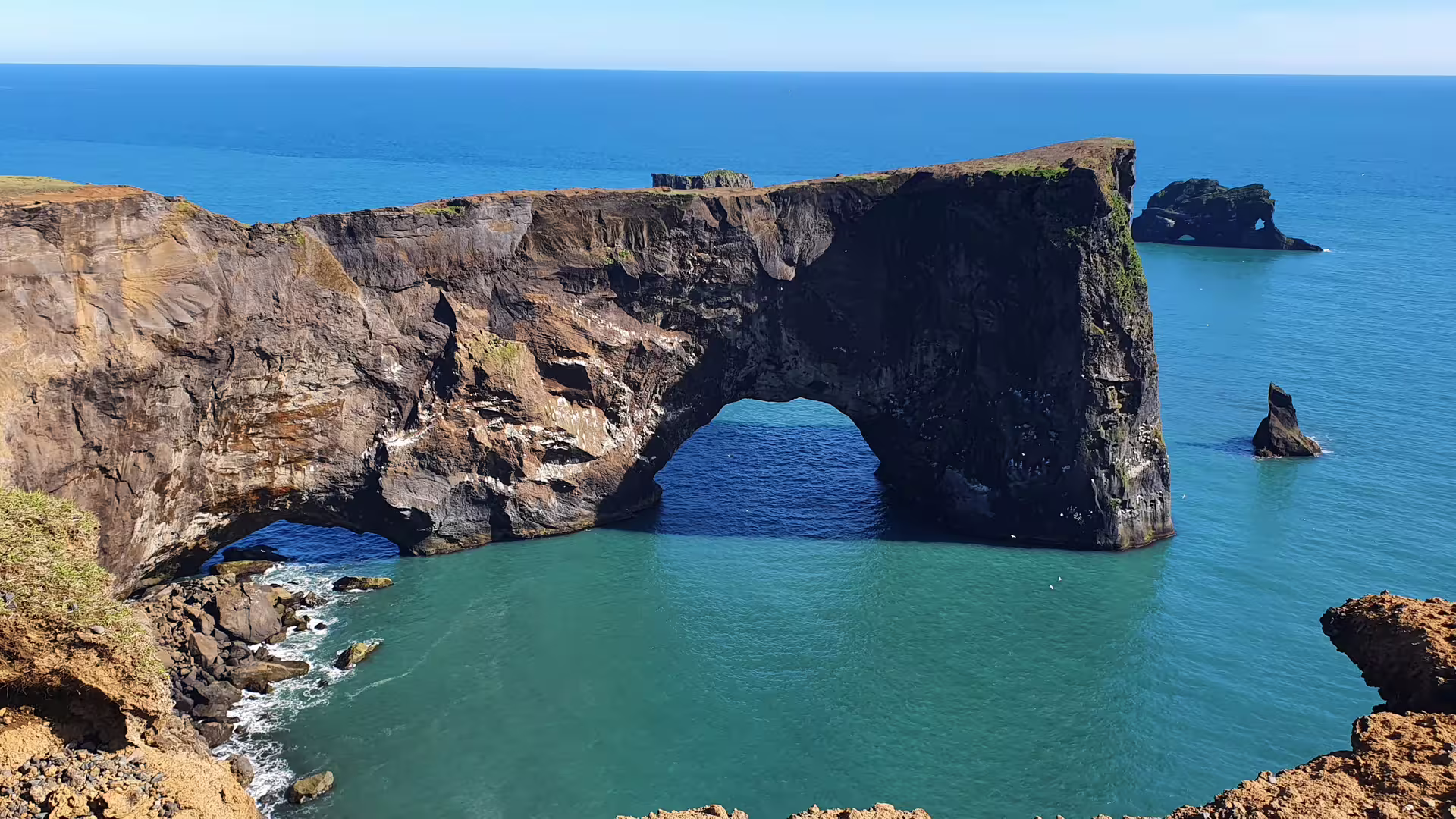 Large natural rock arch over turquoise waters along Iceland's black beach coastline on a sunny day