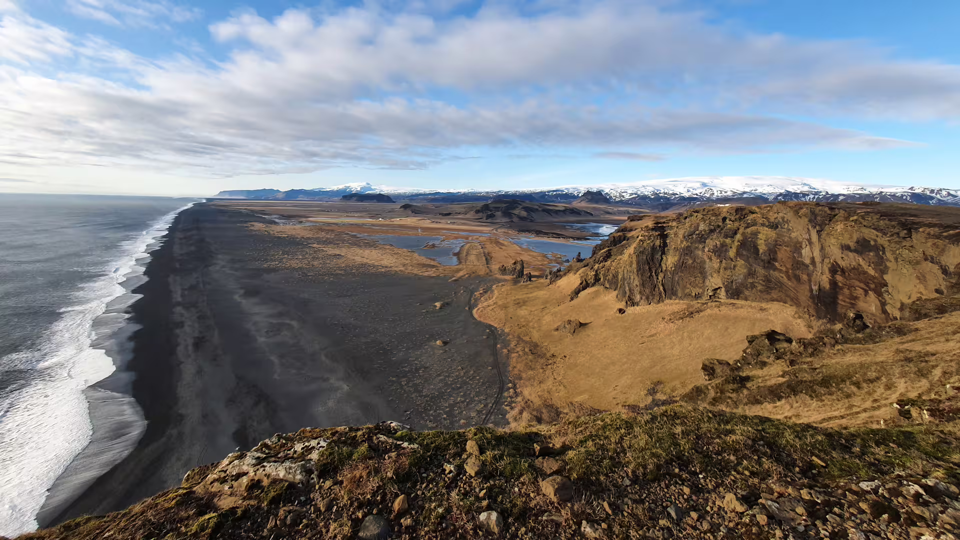Black sand beach stretching along Iceland's south coast with snow-capped mountains and volcanic cliffs