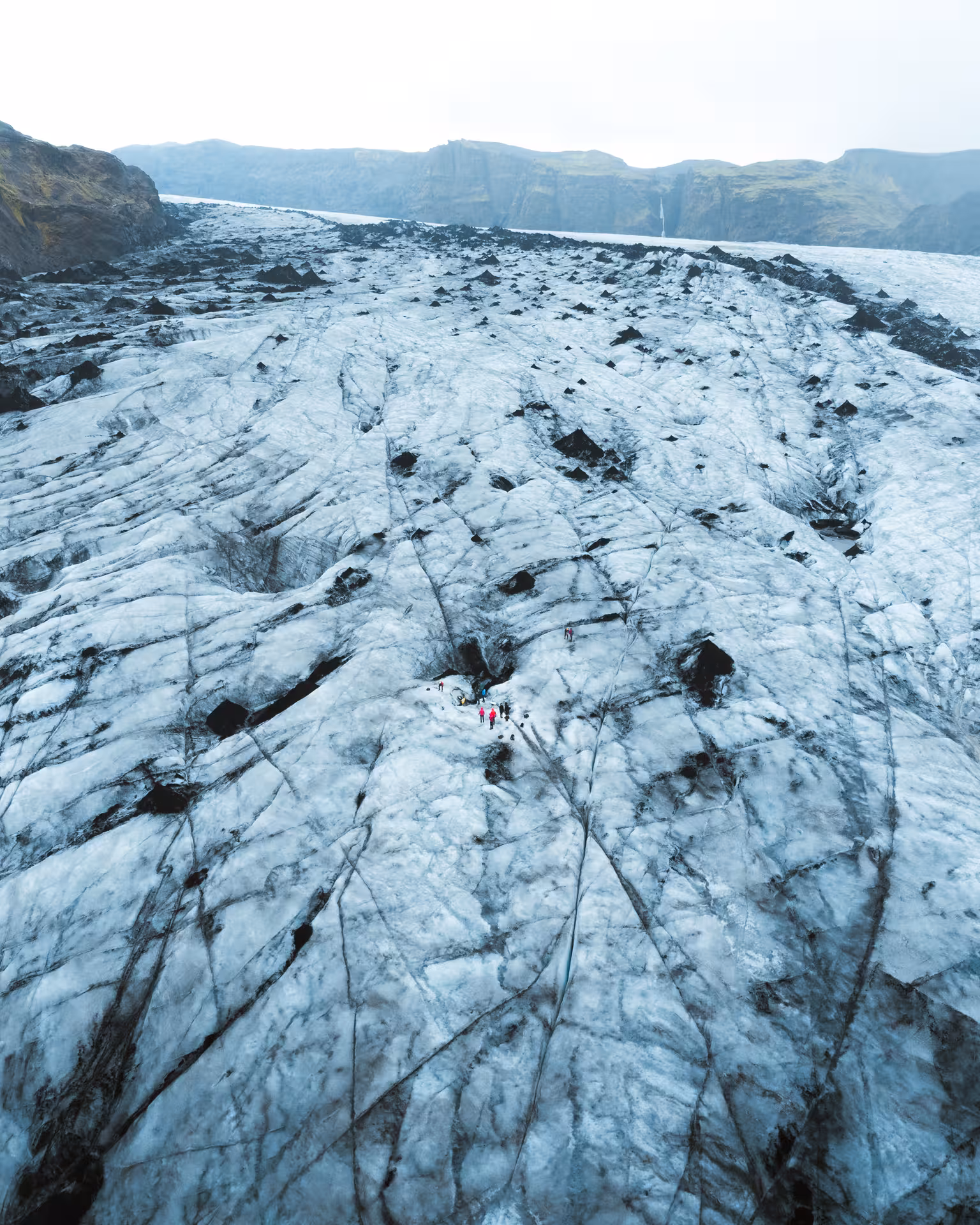 Aerial view of hikers exploring a vast glacier with dark crevasses and rocky outcrops in South Iceland
