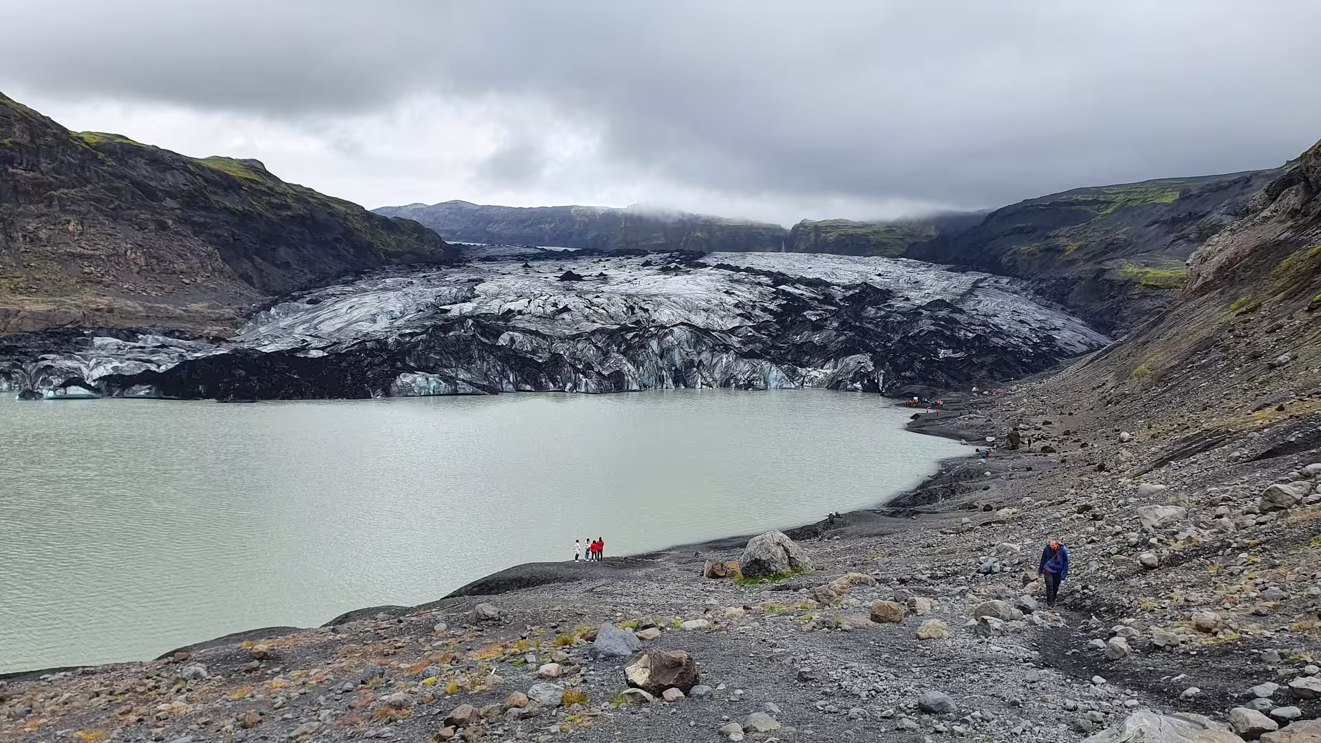 Glacial lagoon with dark ash-covered glacier tongue and tourists on rocky shoreline in South Iceland