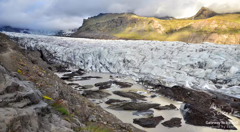 Glacier tongue with crevasses and meltwater pools against green mountains in South Iceland