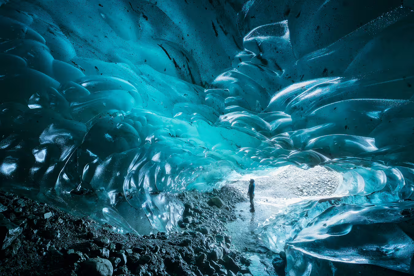 Person standing inside a glowing turquoise glacial ice cave with dramatic sculpted ice walls and rocky floor