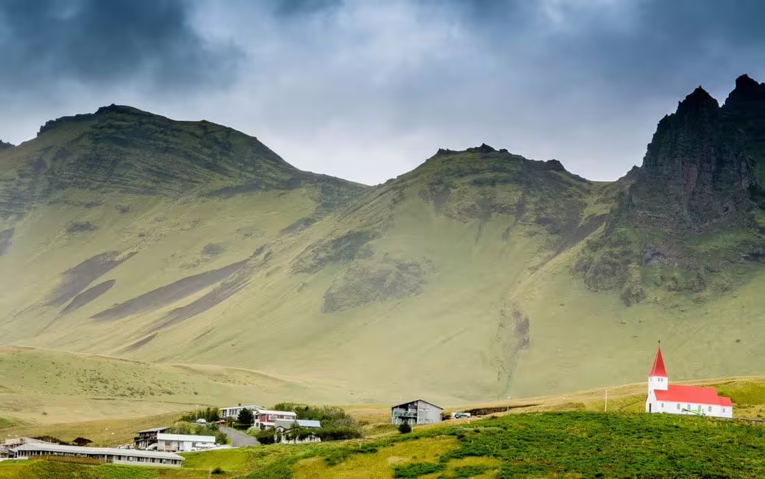 White church with red roof on green hillside below dramatic mountains in South Iceland