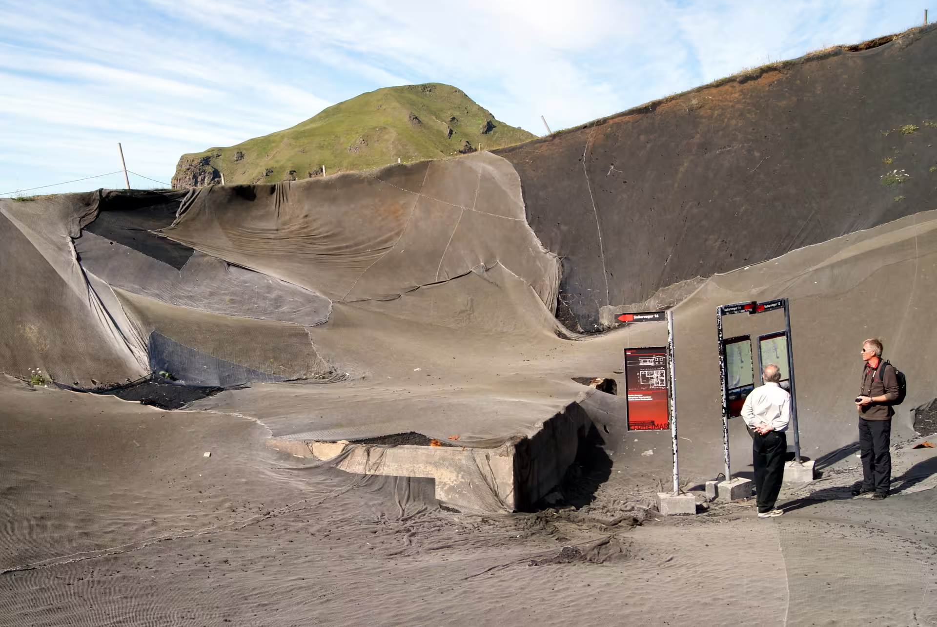 Tourists reading information boards at volcanic ash-covered site in South Iceland with green hills behind