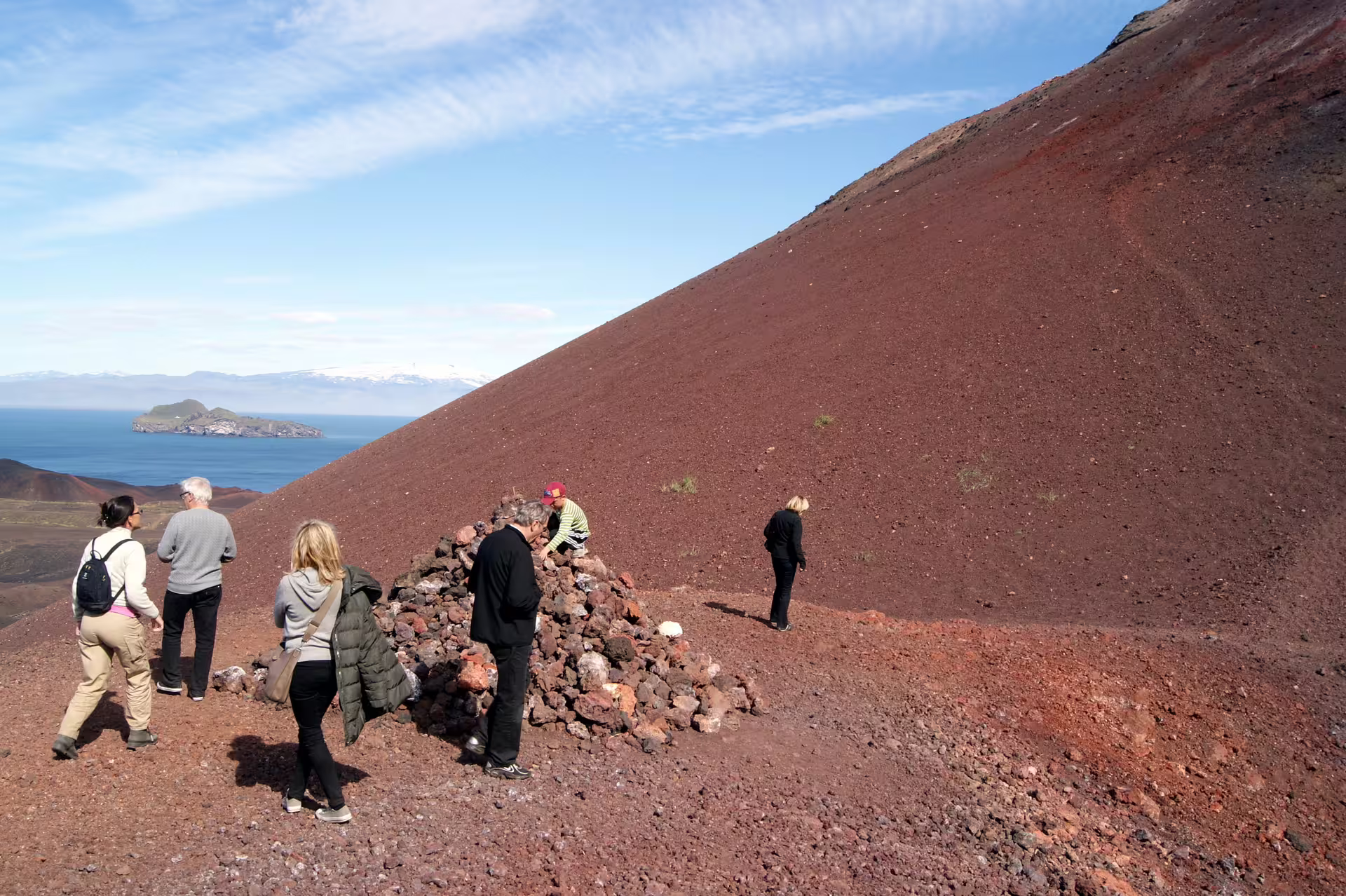 Tourists exploring red volcanic crater on South Iceland tour with ocean and snowy mountains in background