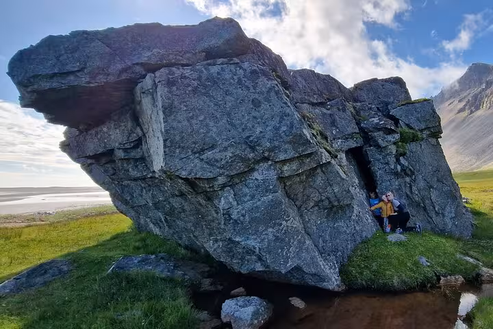 Two people sheltering inside a massive glacial erratic boulder in Southeast Iceland's green landscape