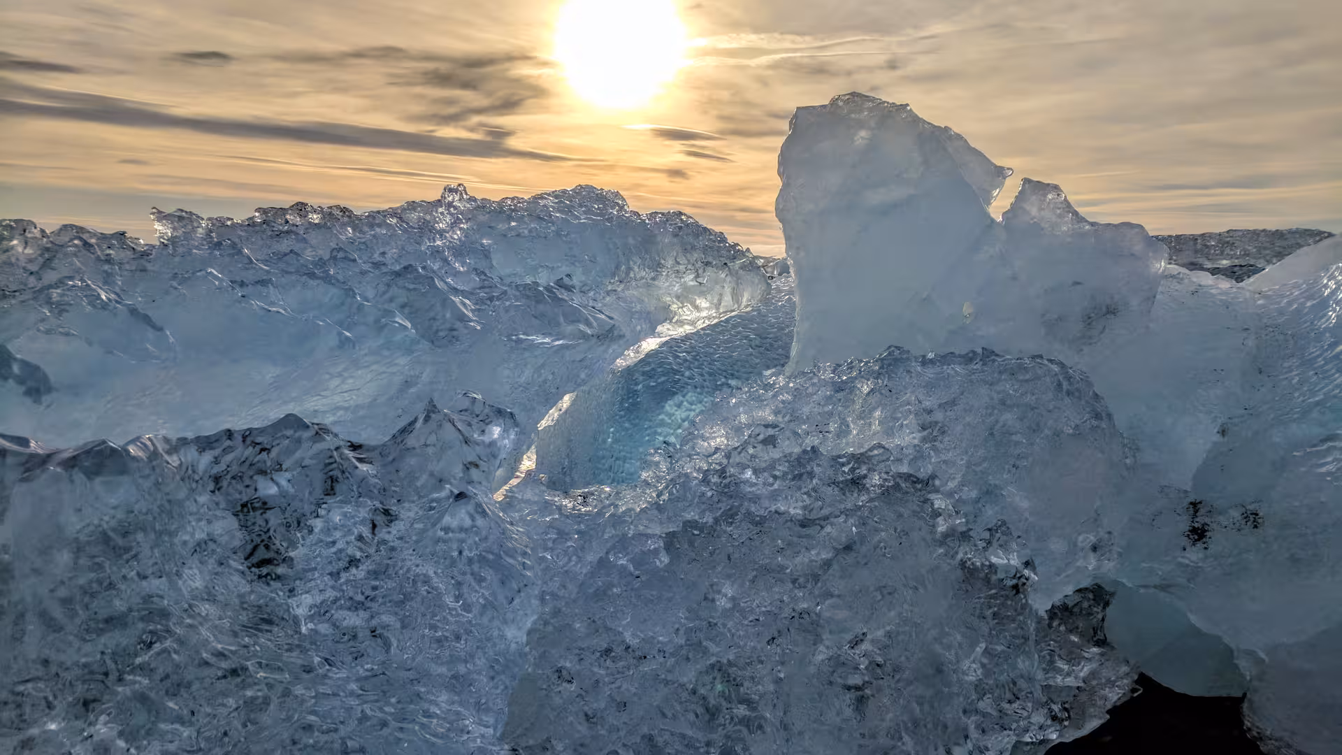 Translucent glacier ice chunks on black sand beach with glowing sun behind clouds in Southeast Iceland