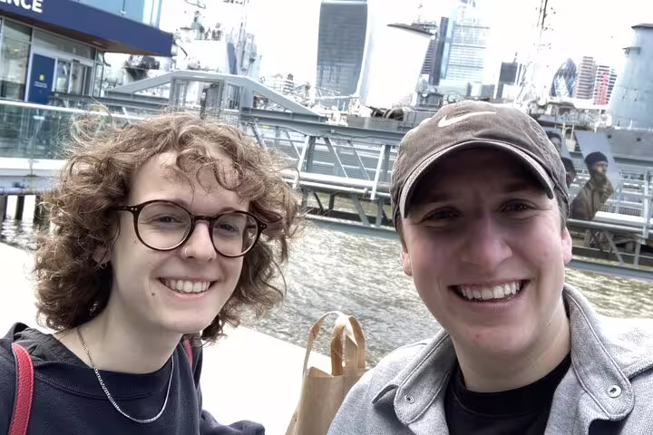 Two smiling visitors posing near the Thames waterfront with a historic warship in background, Southwark London