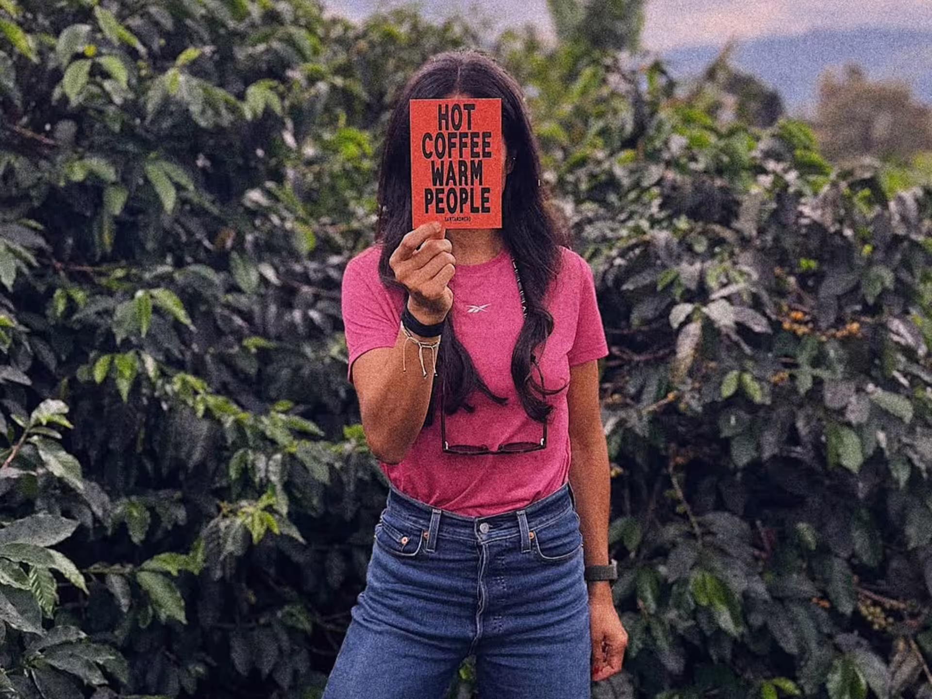 Person holding a sign saying 'Hot Coffee Warm People' in front of lush coffee plants in Turin.