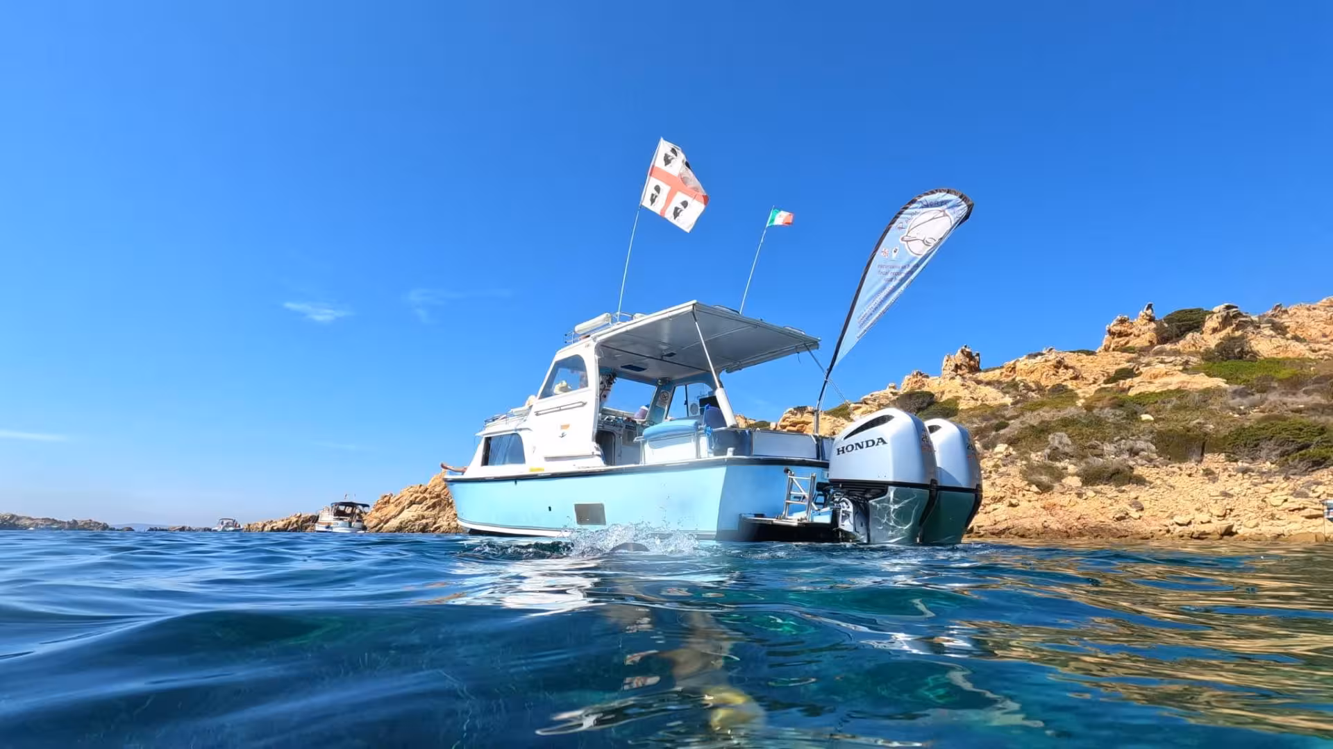 A speedboat with flags anchored by rocky coastline in the clear waters of La Maddalena Archipelago.