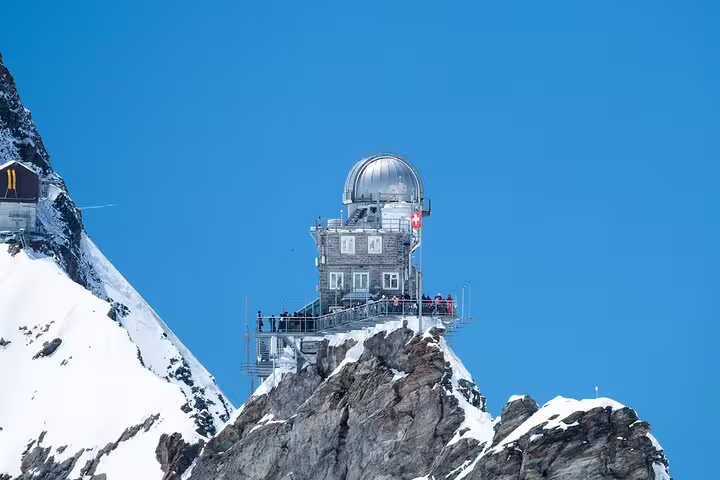 Sphinx Observatory atop Jungfraujoch summit with Swiss flag and snow-covered Alps under blue sky