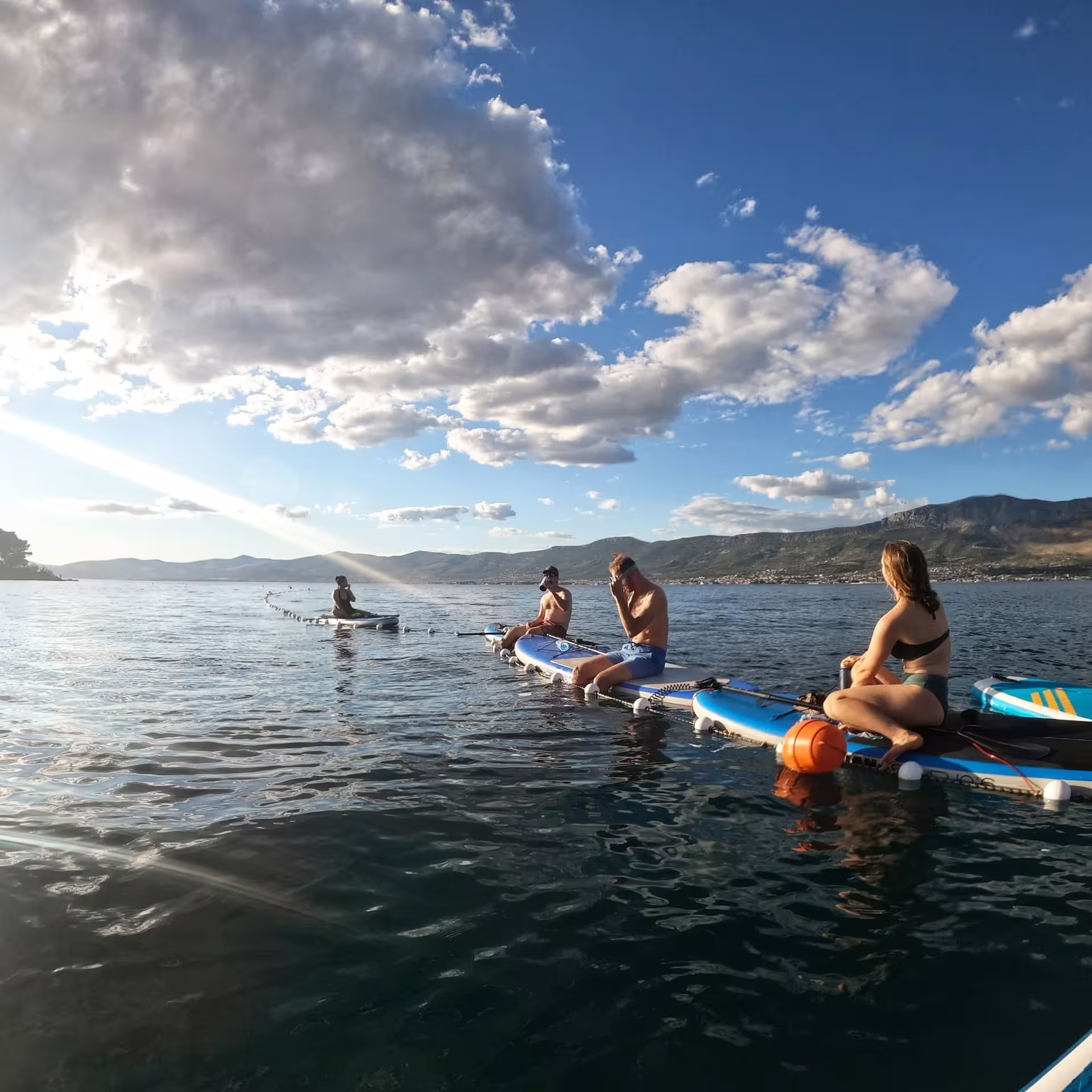 Group resting on paddleboards in Split bay during sunset SUP tour, calm Adriatic sea with mountains and clouds