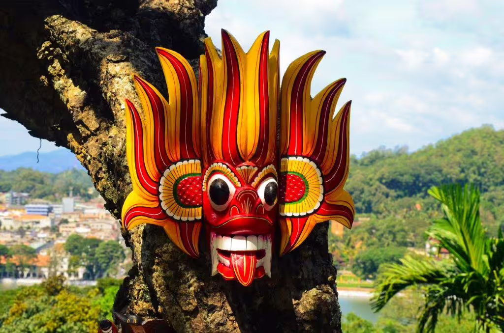 Vibrant traditional Sri Lankan mask with intricate designs hanging on a tree with a scenic backdrop.