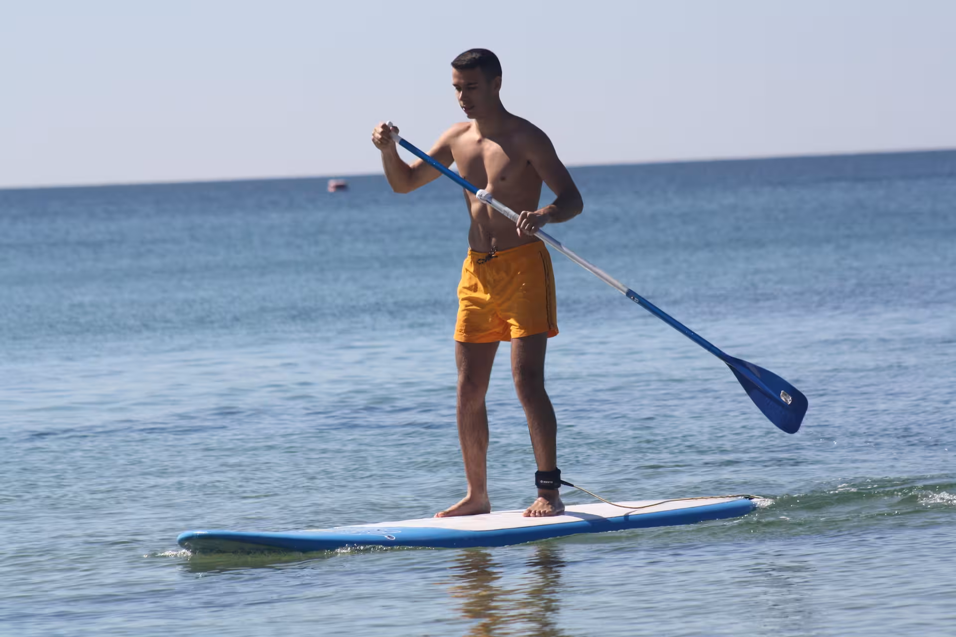 Young man in orange shorts paddling on a stand up paddle board rental on calm blue ocean waters