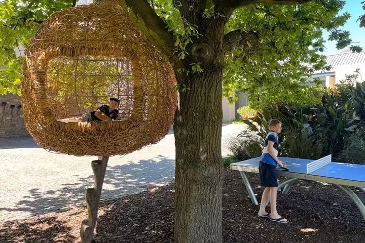 Child relaxes in a woven nest chair while another plays ping pong at a scenic Stellenbosch wine estate.