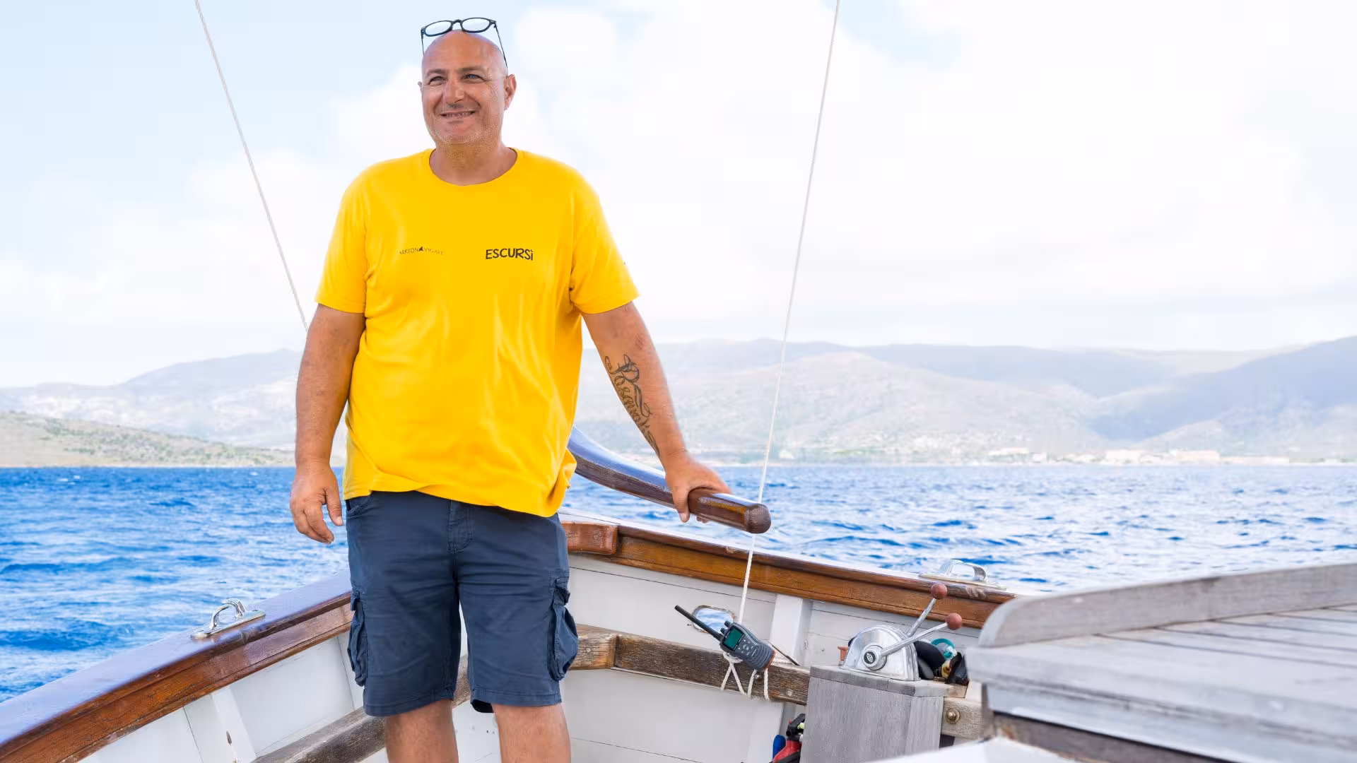 Smiling guide in yellow shirt on a sailing trip from Stintino to Asinara with scenic mountains in the background.