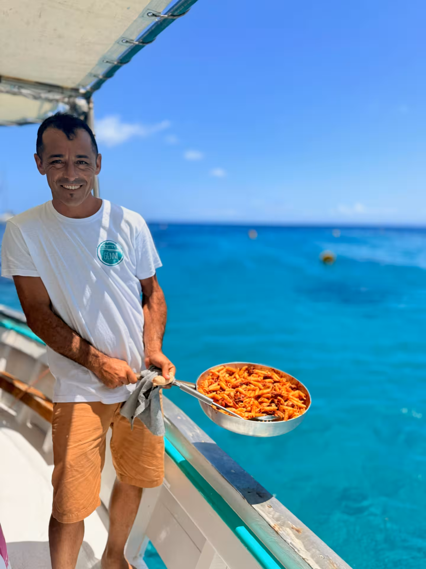 Man presenting a pan of seafood pasta on a boat with vibrant blue waters in Asinara Park.