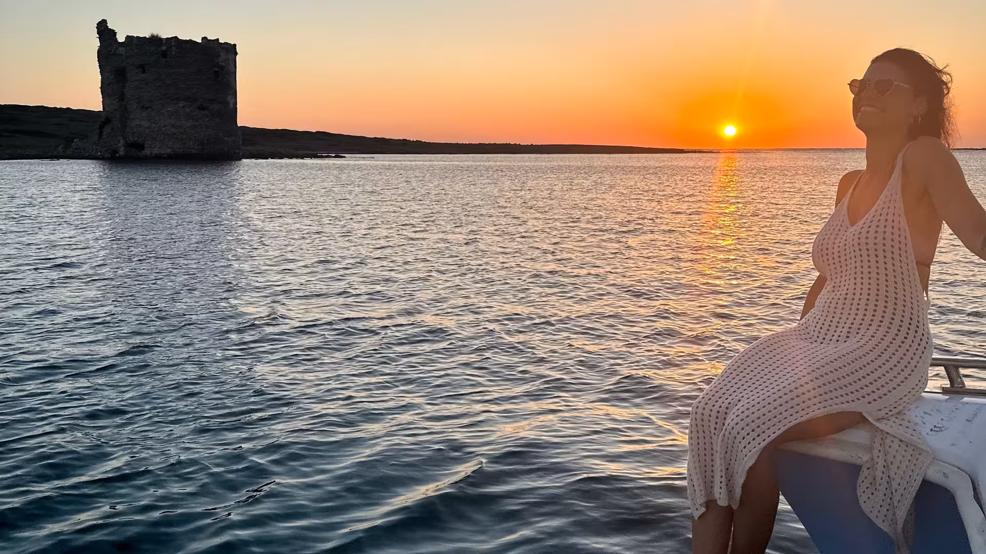 Woman enjoying sunset view from a catamaran in the Gulf of Asinara with ancient tower silhouette.