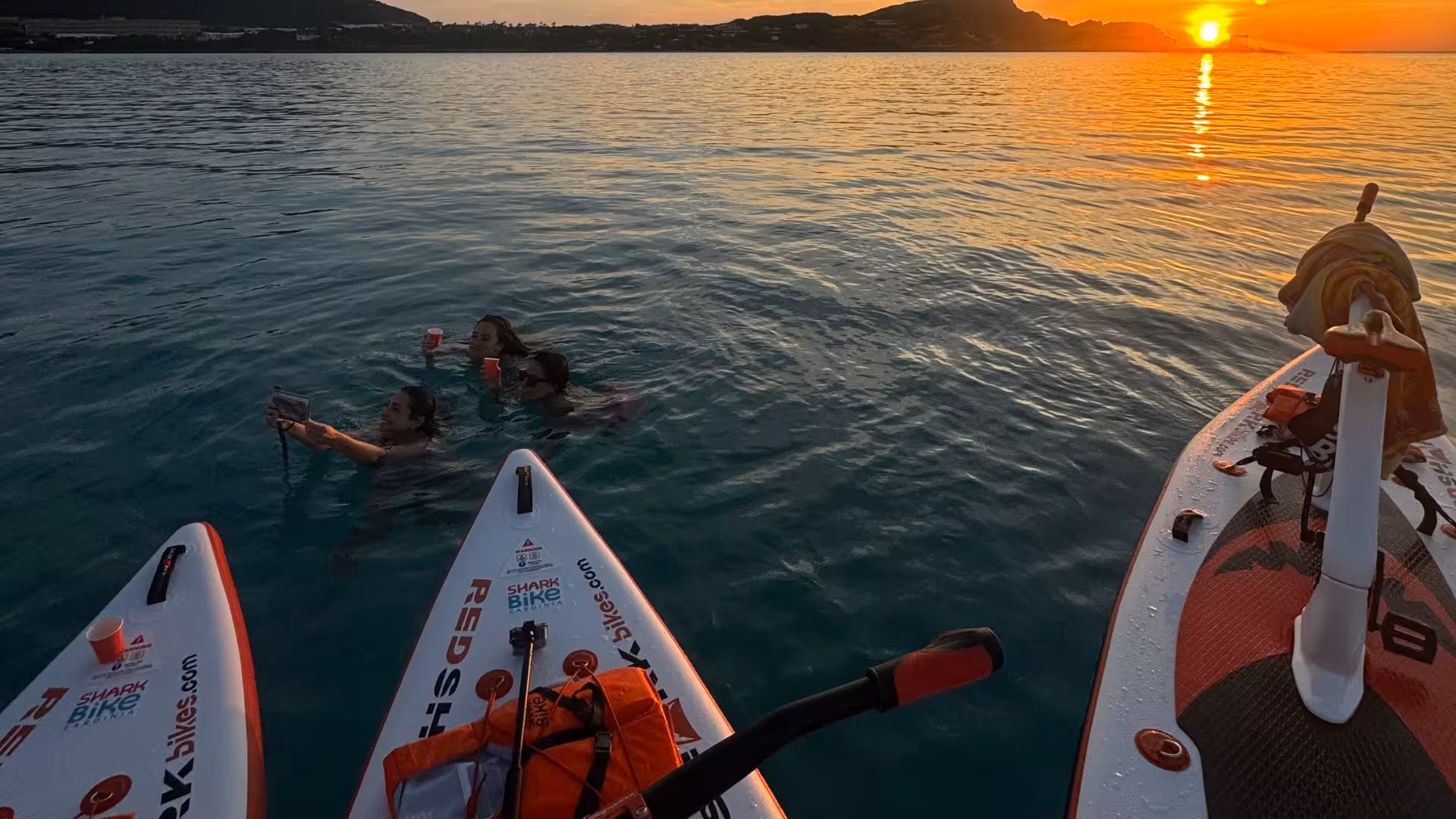 Group of friends swimming near water bikes during a stunning sunset at La Pelosa, Stintino.