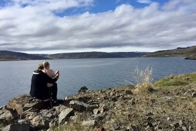 Two people sitting on rocky cliff overlooking a calm fjord with rolling hills in Iceland