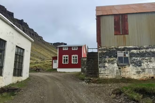 Red Icelandic house on gravel road between old buildings with dramatic mountain backdrop in Strandir