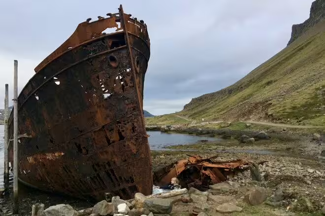 Rusty shipwreck on rocky shore with green Icelandic cliffs and fjord in background