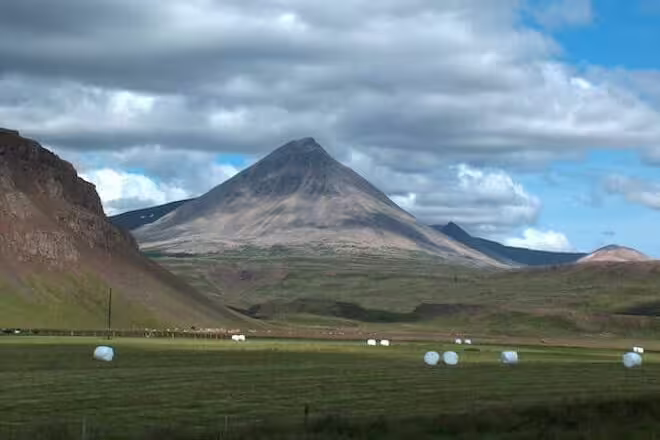 Pyramid-shaped volcanic mountain rising over green valley with white hay bales in Iceland's Strandir region