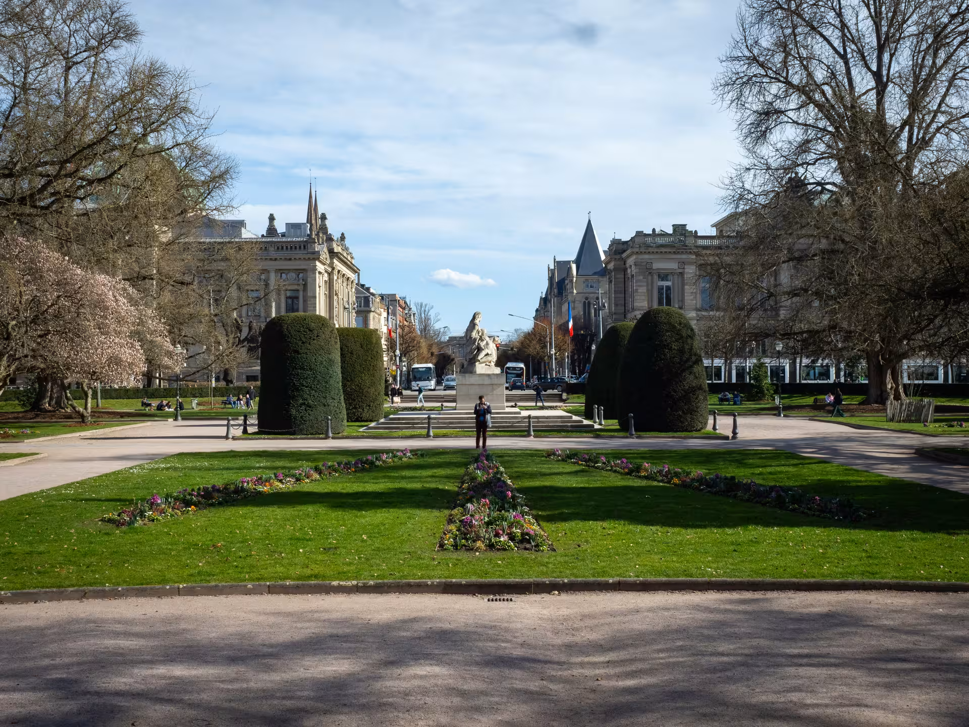 Formal garden with manicured hedges, flower beds, and a statue along a grand boulevard in Strasbourg
