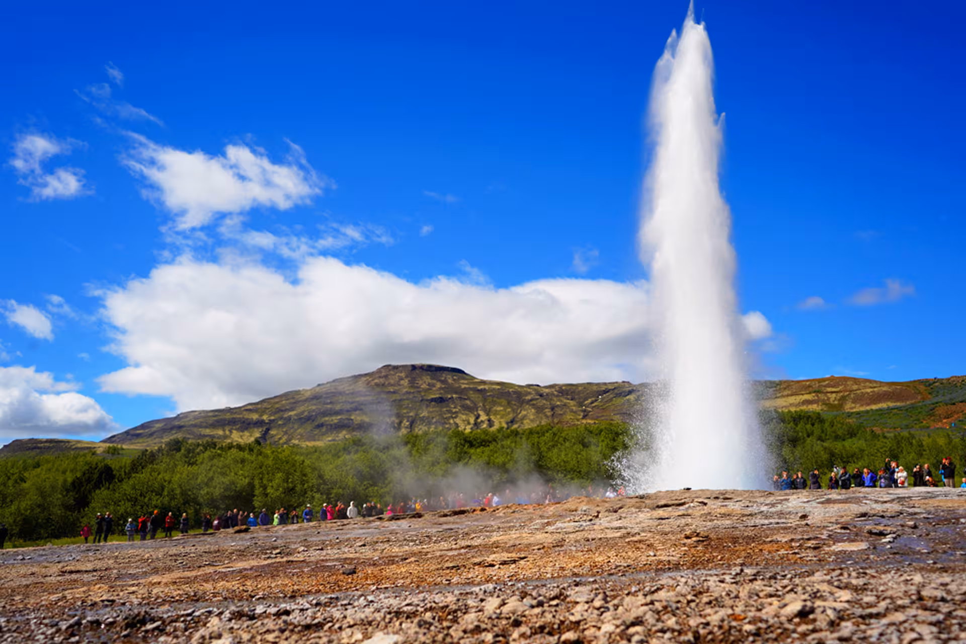 Strokkur geyser erupting on Iceland's Golden Circle with tourists watching against a blue sky