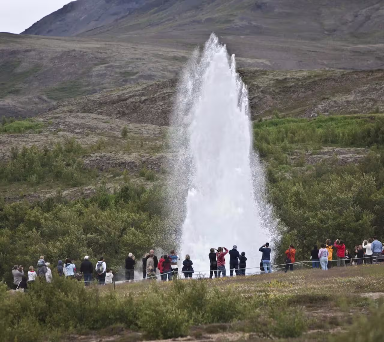 Tourists watching Strokkur geyser erupt in Iceland with green hills in background