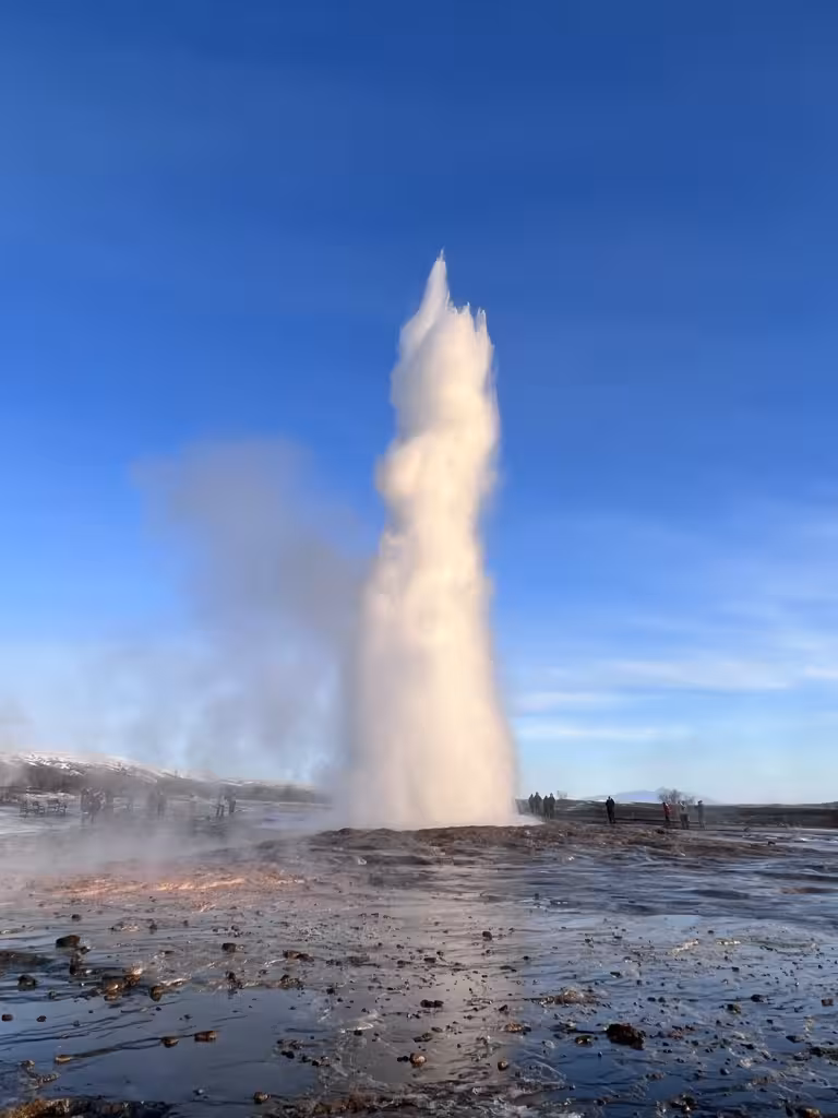 A powerful geyser erupts high into the air at Strokkur, Iceland, surrounded by a scenic geothermal landscape.