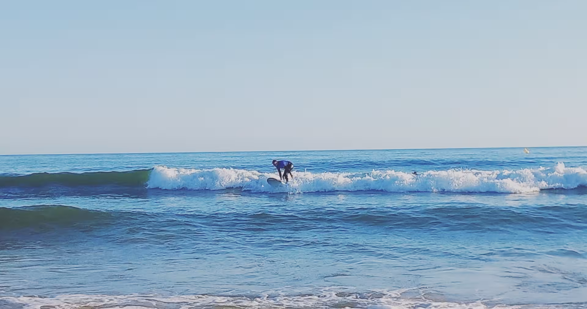 Student catching a small wave in clear blue water on a 3-hour group surf lesson, learning to stand confidently